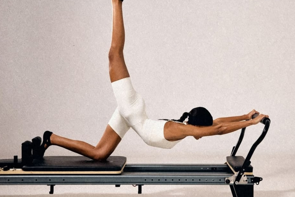 A woman in a white bodysuit performs a Pilates exercise on a reformer machine, with one leg raised high against a plain light gray studio background.